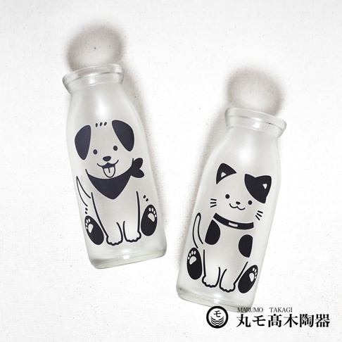 一組兩個透明玻璃牛奶瓶，分別印有可愛的黑白狗狗和貓咪圖案，狗狗戴著領巾，貓咪戴著項圈，瓶身帶有霧面質感。