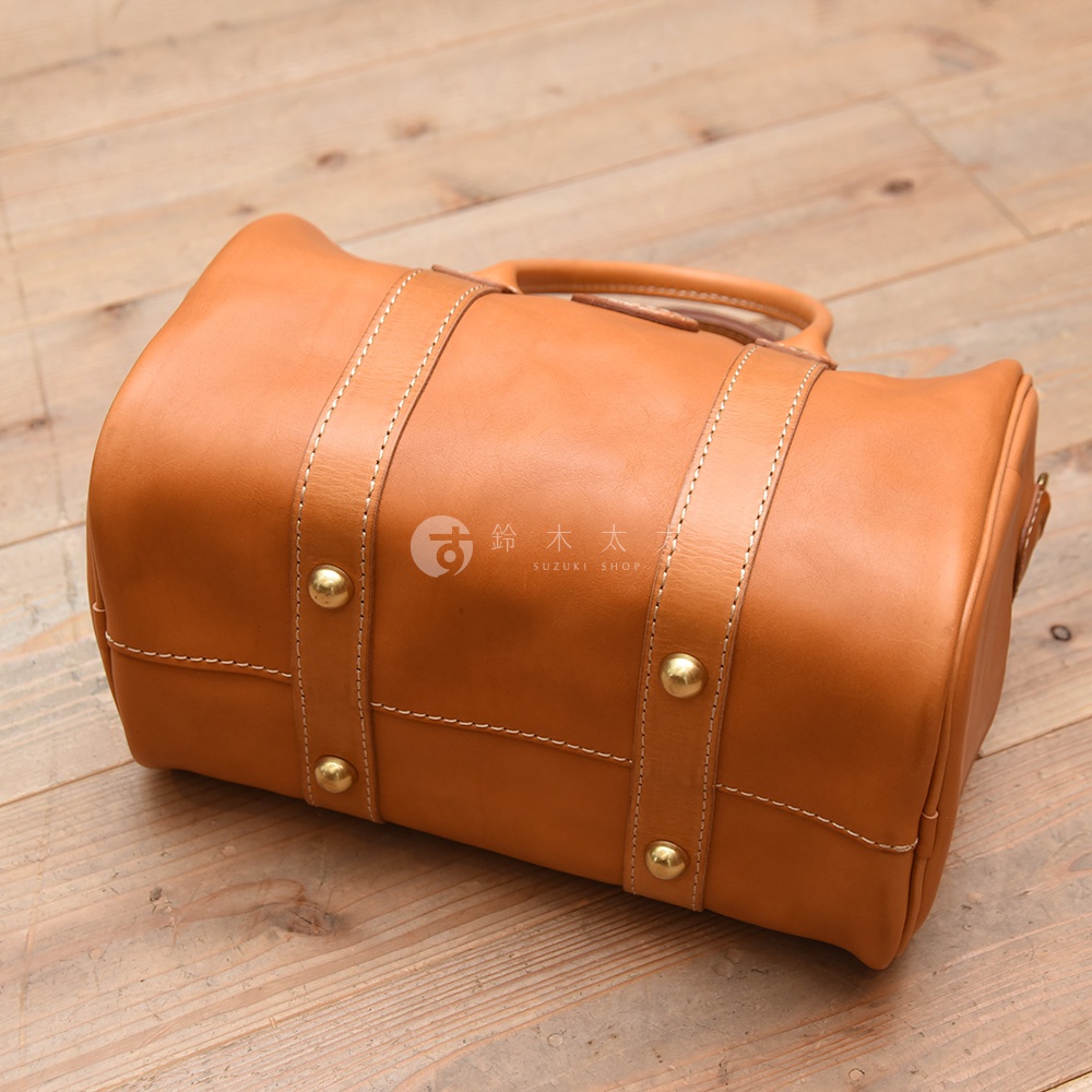 職人手作皮革肩背包，採用天然棕色皮革，搭配銅色金屬卯釘及白色縫線，提把設計，風格簡潔。
