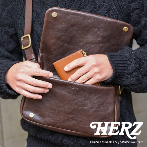 職人手作皮革斜背包，深棕色，附有可調整的肩帶和金屬扣環。包包開口處可見一只手拿出一個淺棕色短夾。