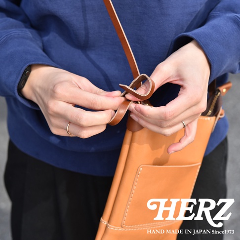 職人手作皮革直式隨身單肩包，採用棕褐色皮革製成，結構簡潔，配有肩帶。包身外側有縫線裝飾的口袋，並有品牌標誌。