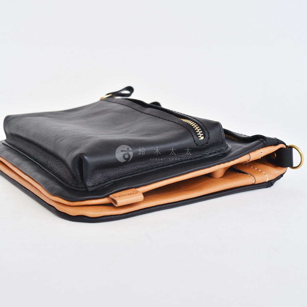 職人手作皮革翻轉雙色直式斜背包，包身採用黑色光滑皮革，翻蓋內側為原色皮革，搭配金色拉鍊與金屬 D 型環。