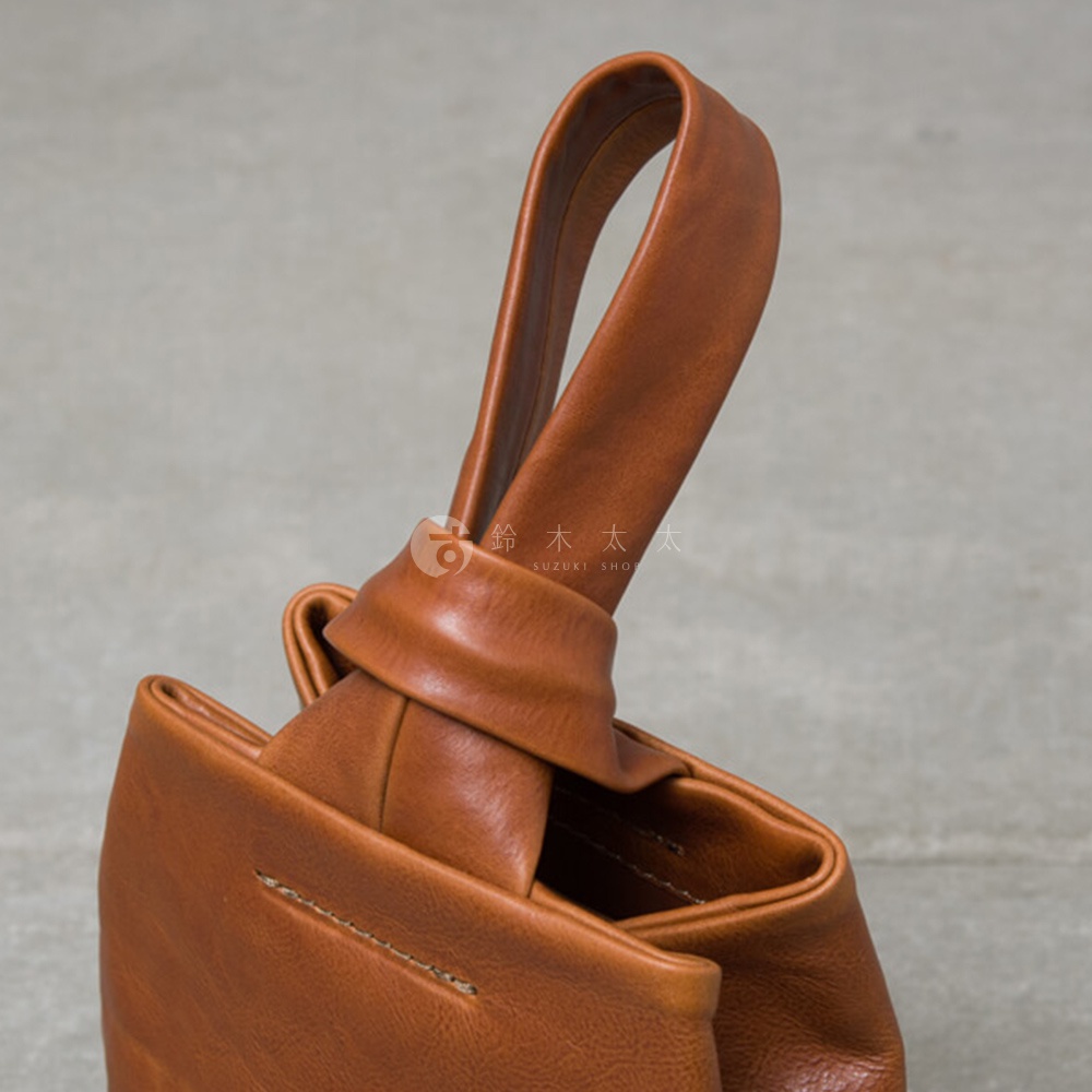 職人手作的棕色皮革單柄手提包，包體結構呈現摺疊設計，提把以皮革繩結造型貫穿，材質為光滑皮革，縫線細緻。