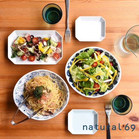 白色八角形餐盤盛裝義大利麵，盤邊有藍色線條圖案。旁邊的圓形藍紋餐盤盛有沙拉，內含萵苣、番茄、黃椒和起司。另有長方形白色餐盤盛裝章魚、番茄、黃瓜與洋蔥沙拉。畫面中有綠色玻璃杯、銀色叉子與夾子。