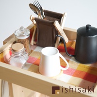 木質收納推車上擺放著一個白色的馬克杯、一個黑色細嘴茶壺、兩個玻璃罐（分別裝有淺褐色顆粒和白色粉末）、一個展示餐具（湯匙和叉子）的木質帆布架，以及一塊橘黃色格紋布。