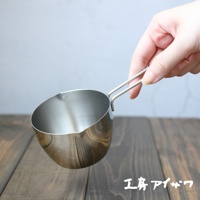 一只手握著一個不鏽鋼長柄量杯，杯身呈圓弧形，帶有傾倒口，表面呈現金屬光澤。