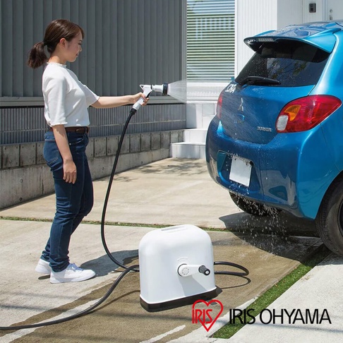 女性使用白色噴槍連接黑色水管，正在清洗藍色汽車的後輪。前方地面放置著一個白色圓角方形水管收納捲筒，側面有黑色捲軸。