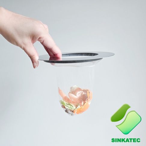 一隻手正拎著一個 SINKATEC 品牌的廚房水槽磁吸濾網，細網袋內裝有剩餘的蔬果殘渣，外圈搭配金屬邊框，背景為簡約淺灰色。