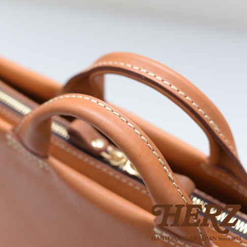 日本HERZ職人手作皮革公事包，近拍皮革提把與縫線細節，右下角為品牌Logo及「HAND MADE IN JAPAN Since1973」字樣。