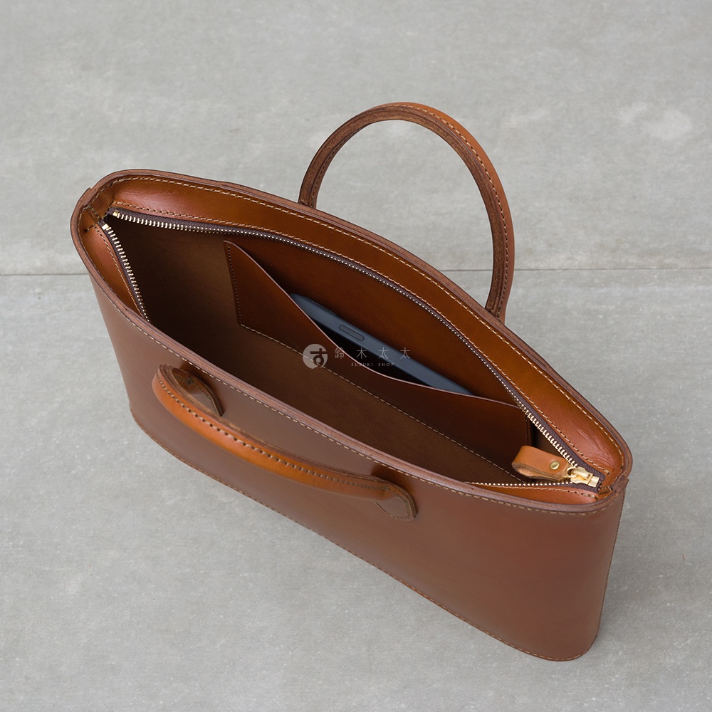 職人手作皮革Organ薄型公事包，以光滑棕色皮革製成，設有頂部提把與拉鍊開口，內部可見手機放置夾層。