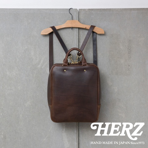 一個深棕色皮革後背包，掛在木製衣架上，背包有手柄和肩帶，背景是灰色牆壁，右下角有HERZ品牌標誌和HAND MADE IN JAPAN Since1973字樣